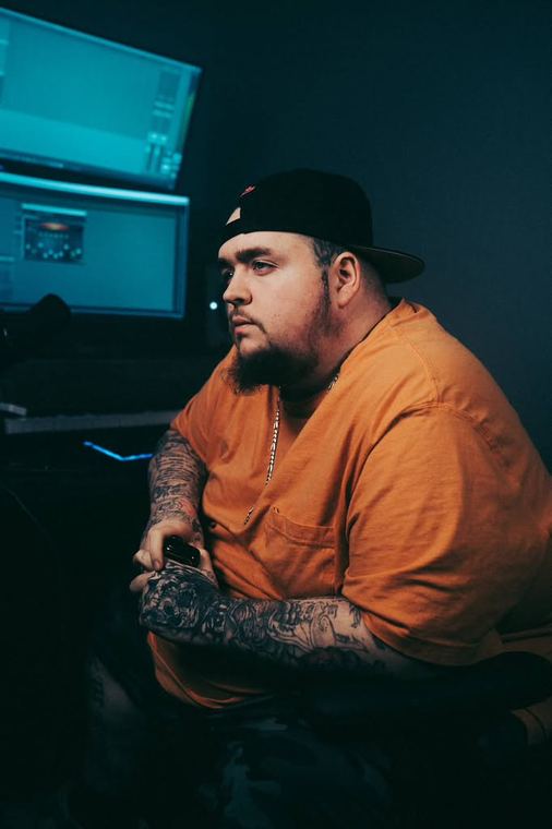 Seated at the studio desk in an orange tee